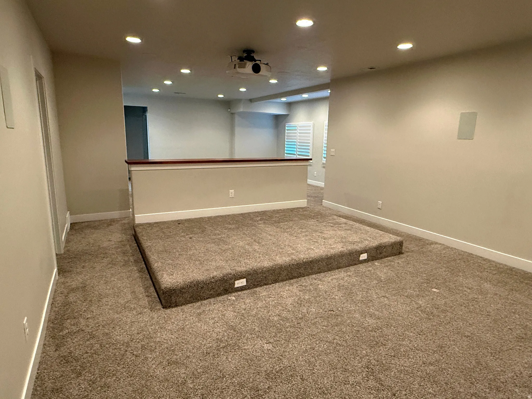 Carpeted media room featuring a raised platform, recessed lighting, ceiling-mounted projector, built-in speakers, and a half-wall partition with a wood cap