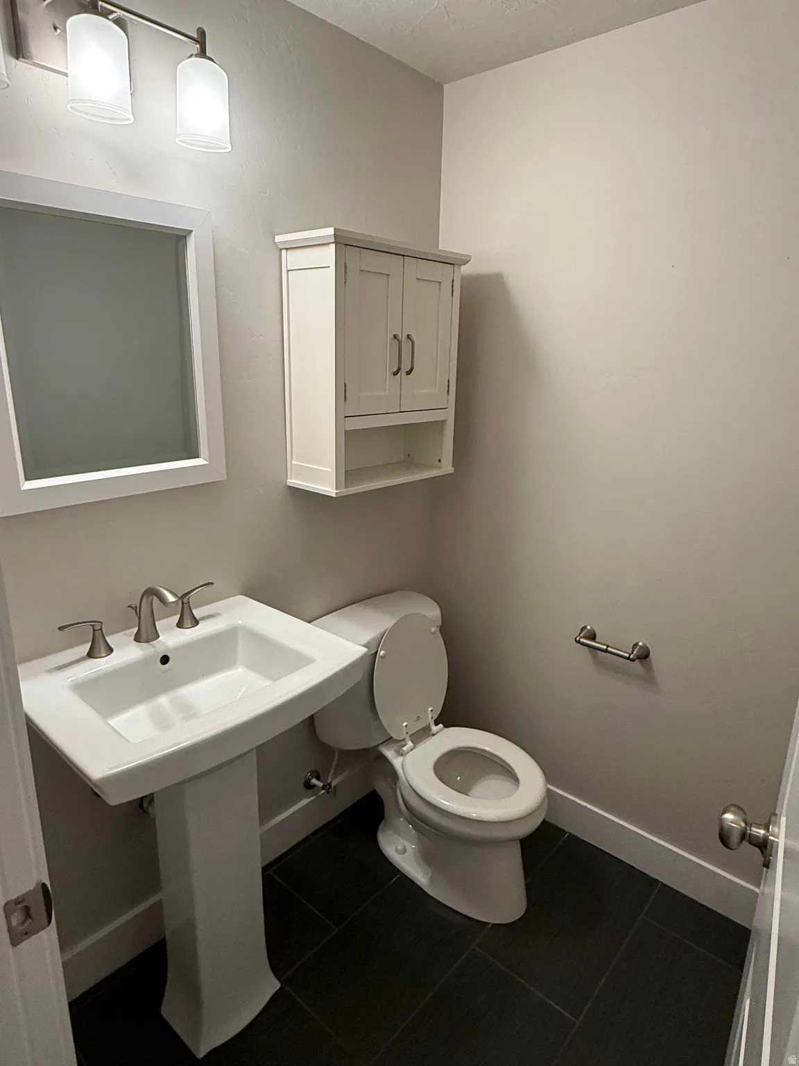 Powder room featuring a pedestal sink with brushed nickel fixtures, a white framed mirror, and dark tile flooring