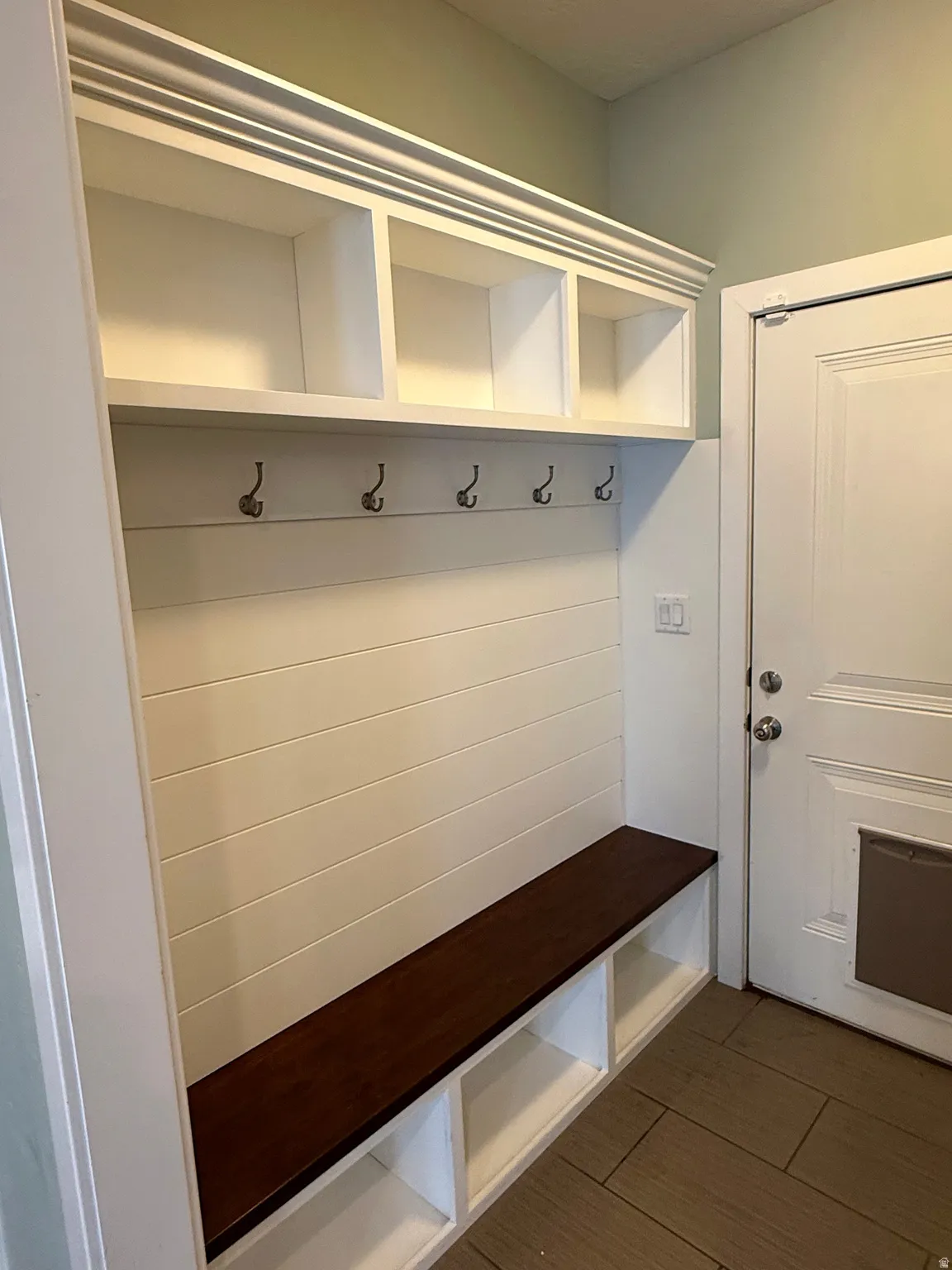 Built-in white storage bench with a dark wood-finish seat, shiplap-style back panel, five coat hooks, and upper cubby shelving with crown molding