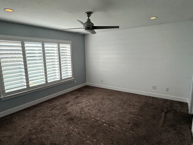 Spacious room featuring a white shiplap accent wall, white plantation shutters, a modern ceiling fan, recessed lighting, and soft brown carpet