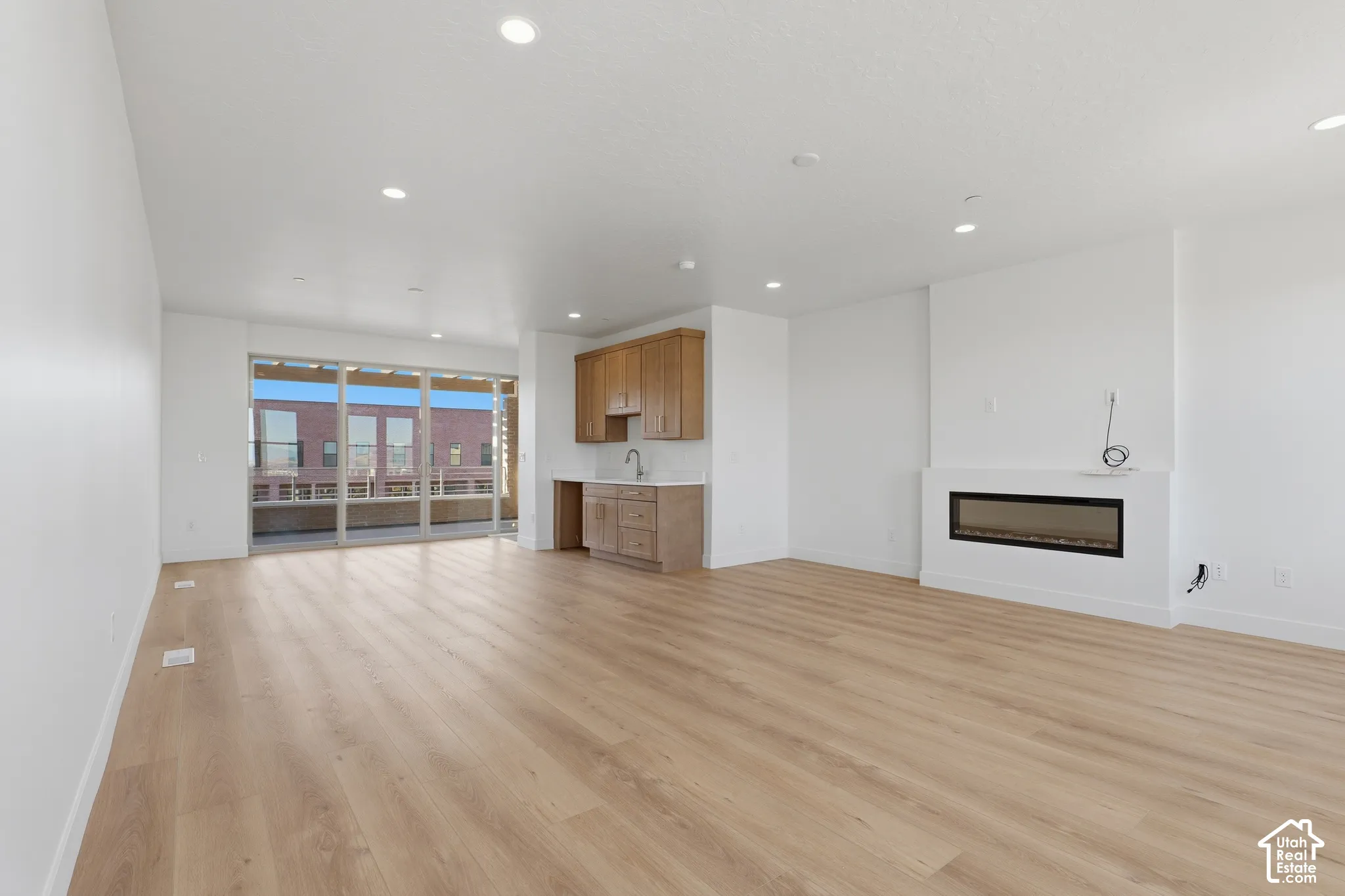 Open-concept interior featuring light-tone wood-finish flooring, recessed lighting, a linear built-in fireplace, natural wood cabinetry, and floor-to-ceiling glass sliding doors