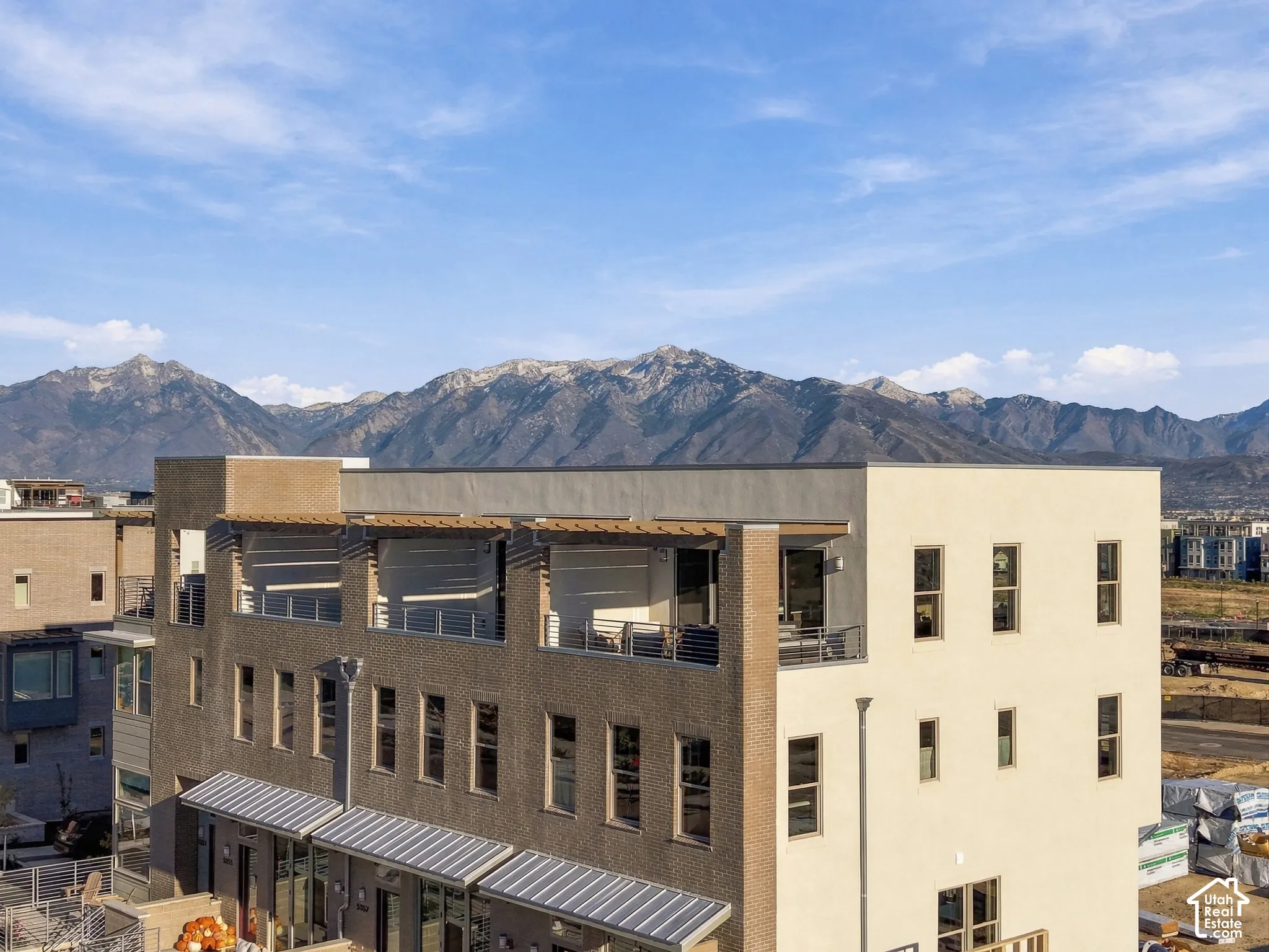 Multi-level exterior featuring a brick and stucco facade, top-floor balcony with horizontal railing, metal awnings, and mountain backdrop