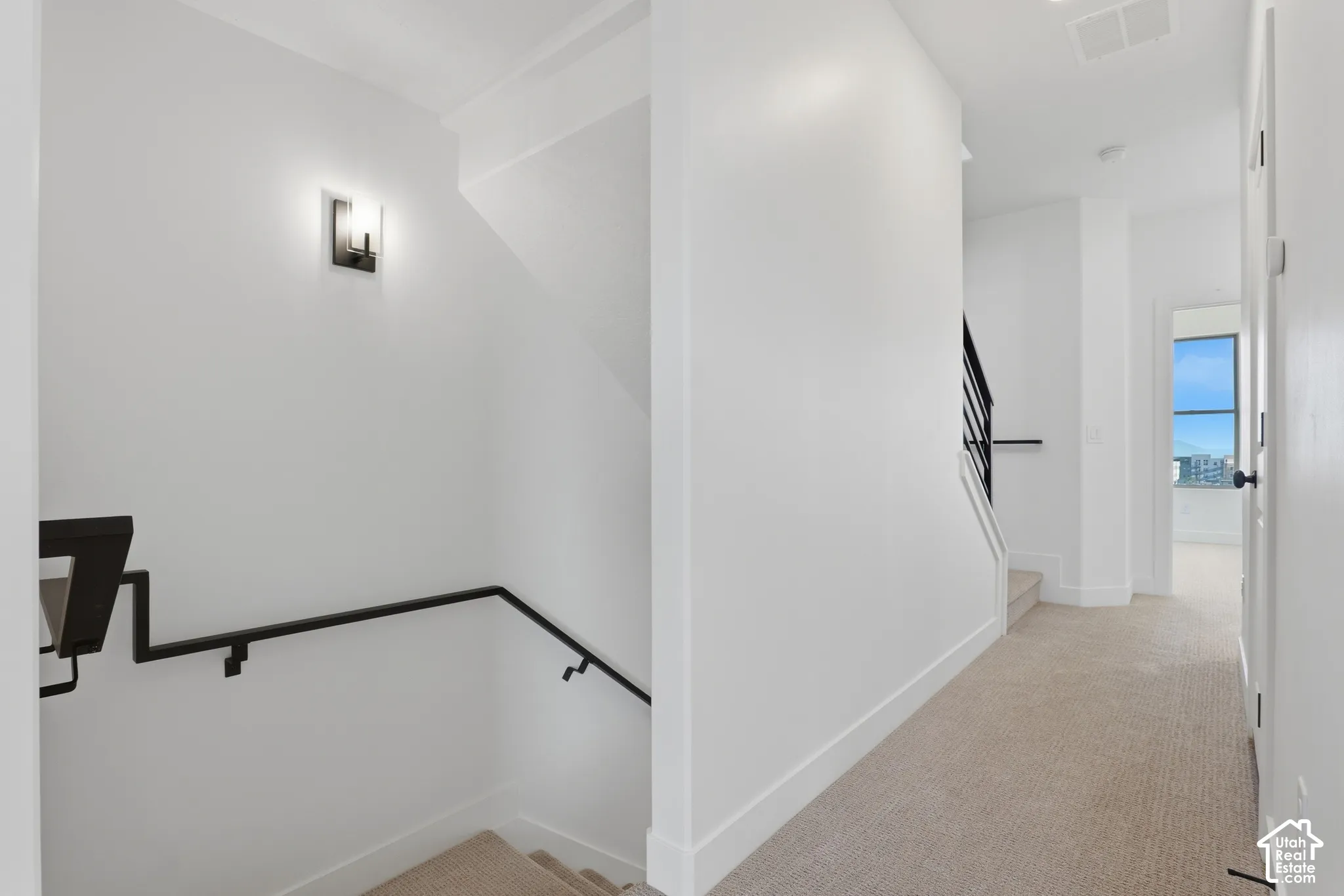 Neutral-toned carpeting, minimalist wall-mounted lighting fixture, and a staircase featuring a matte black metal railing