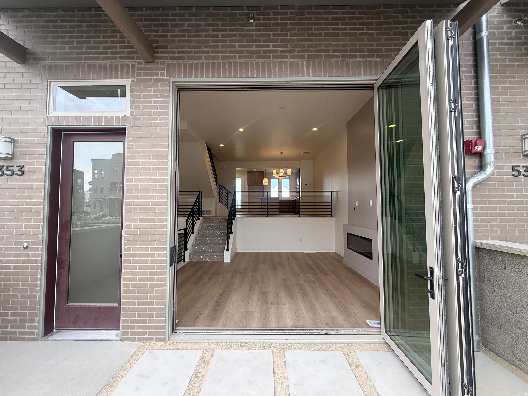 Expansive bi-fold glass doors open to a contemporary interior featuring wood-finish flooring, a linear fireplace, and a modern staircase with dark metal railings