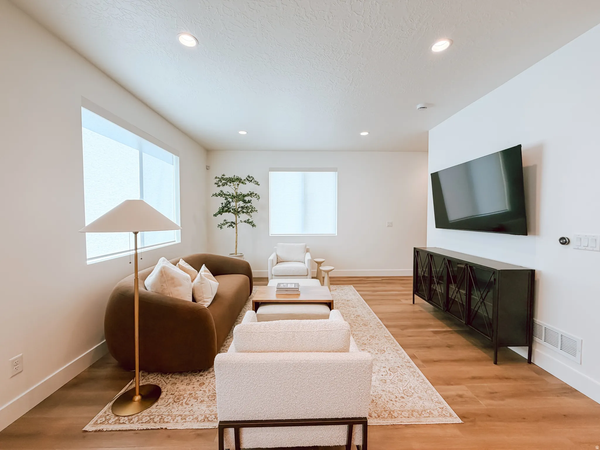 Spacious living area featuring bright white walls, recessed ceiling lighting, wood-finish flooring, and multiple windows with roller shades