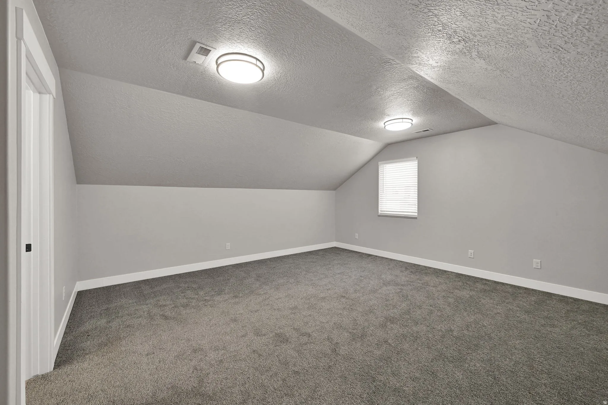 Sloped ceiling attic room with gray carpet, white baseboards, a single window with blinds, and flush-mount lighting fixtures