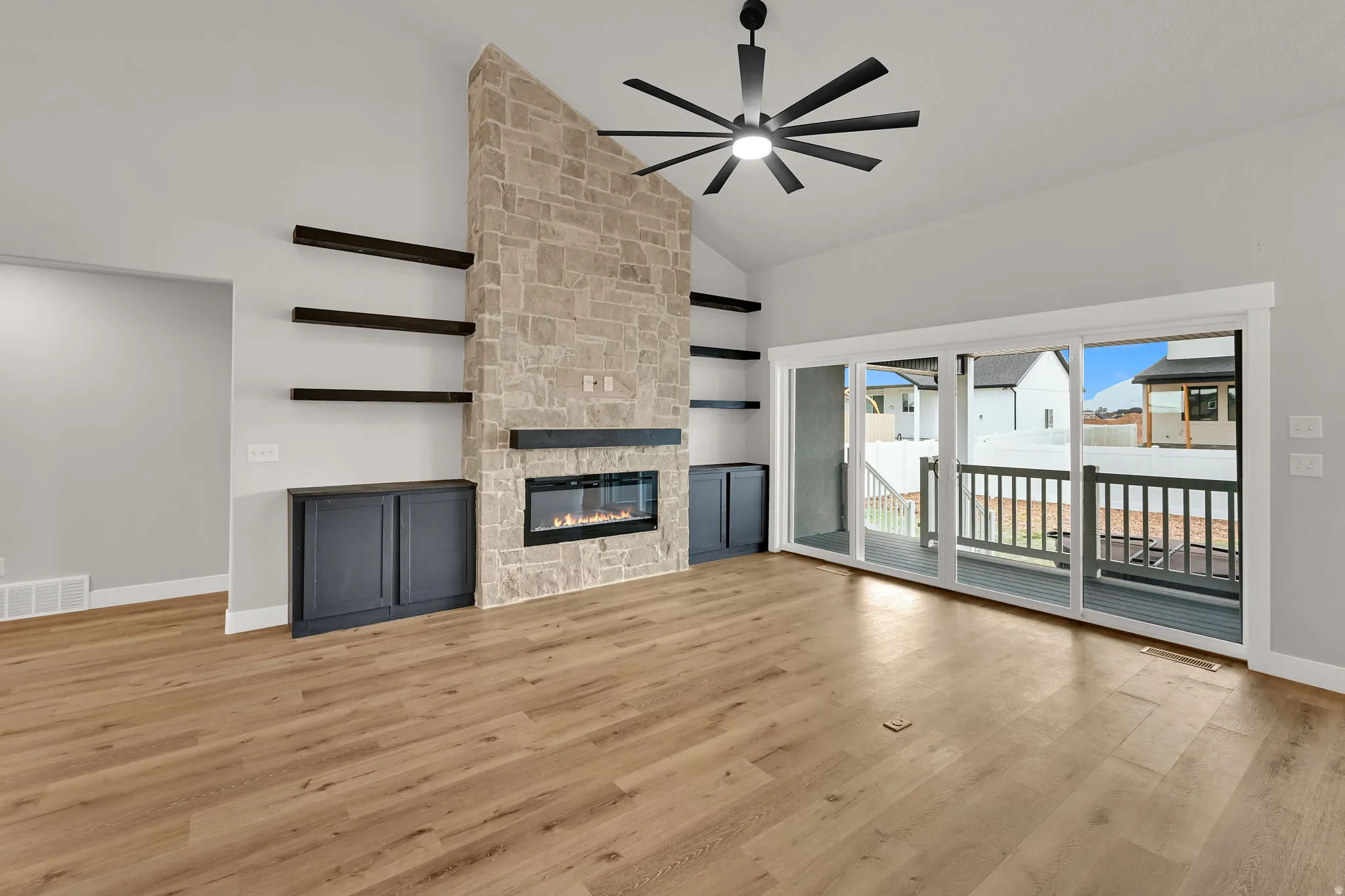 Stone-clad fireplace with built-in electric insert, wood-finish flooring, vaulted ceiling with a modern ceiling fan, and sliding glass doors leading to a deck