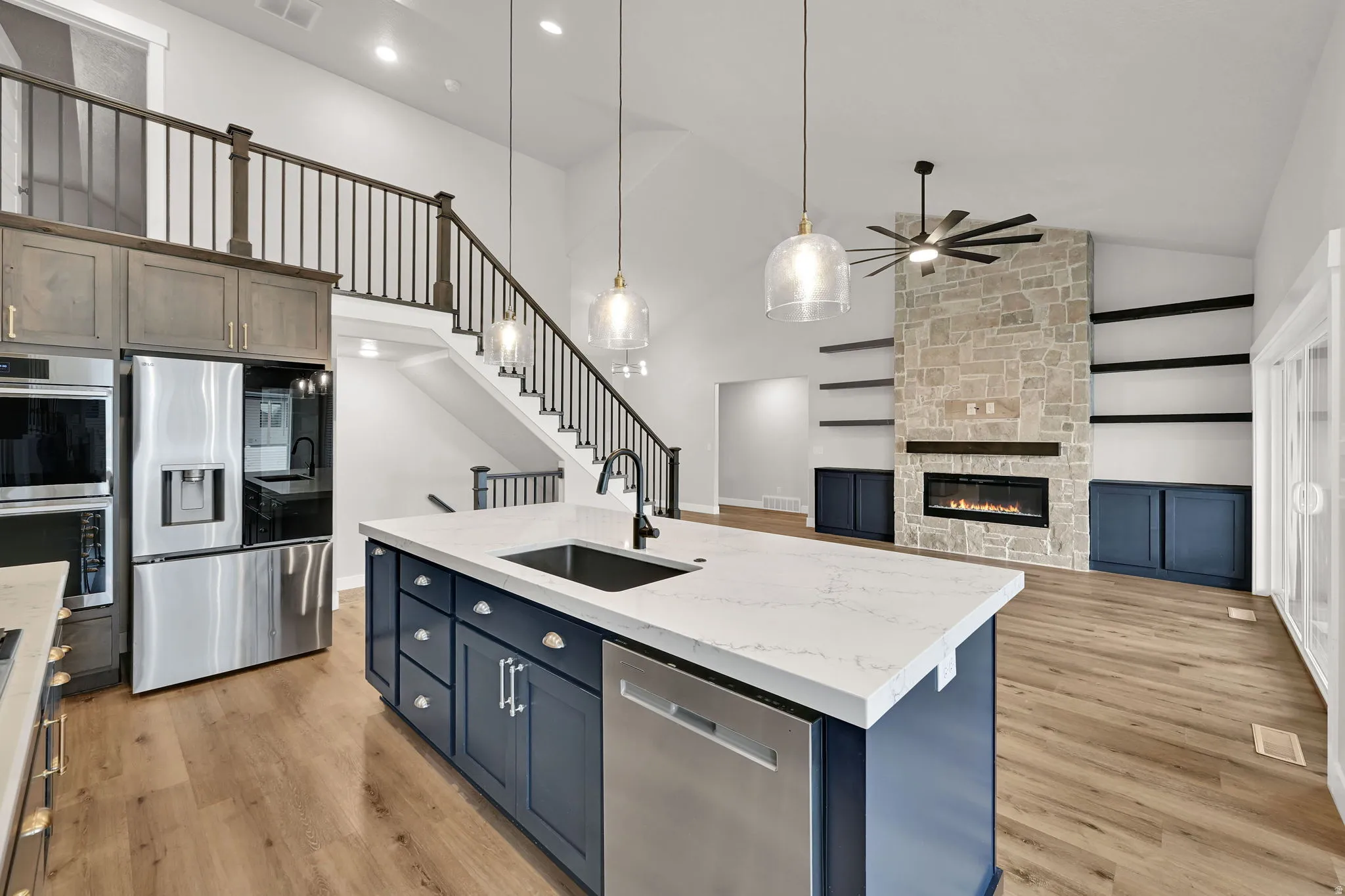 Vaulted ceiling with an integrated ceiling fan, stone-faced fireplace with a mantel, built-in shelving, wood-finish flooring, and an island with a sink and pendant lighting