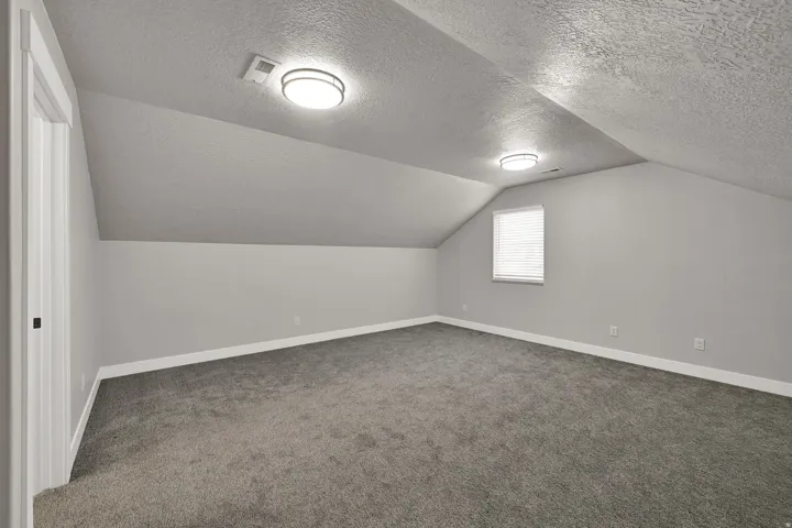 Sloped ceiling attic room with gray carpet, white baseboards, a single window with blinds, and flush-mount lighting fixtures