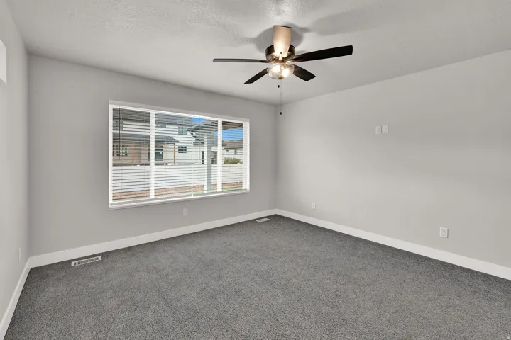 Expansive room with neutral gray carpeting, a large window with white blinds, and a ceiling fan with integrated lighting