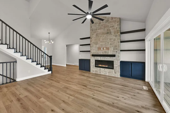 Vaulted ceiling with an integrated ceiling fan, stone-clad fireplace, built-in shelving and cabinetry, wood-finish flooring, and sliding glass door