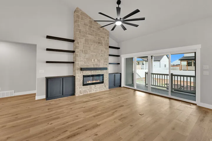 Stone-clad fireplace with built-in electric insert, wood-finish flooring, vaulted ceiling with a modern ceiling fan, and sliding glass doors leading to a deck