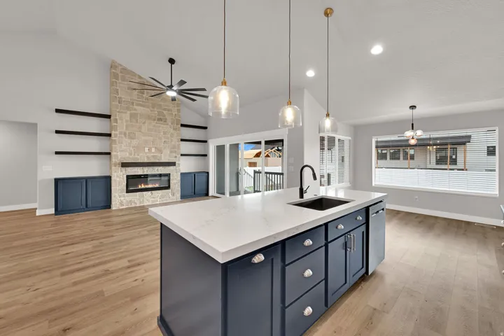 Vaulted ceilings, wood-finish flooring, stone-clad fireplace with built-in shelving, kitchen island with integrated sink, and recessed lighting