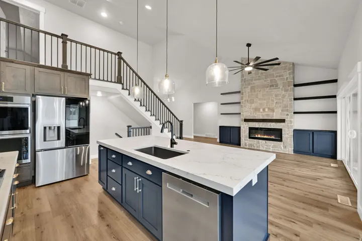Vaulted ceiling with an integrated ceiling fan, stone-faced fireplace with a mantel, built-in shelving, wood-finish flooring, and an island with a sink and pendant lighting