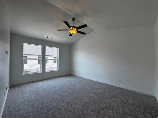 Spacious room featuring vaulted ceilings, a ceiling fan with integrated lighting, two prominent windows, and wall-to-wall carpeting
