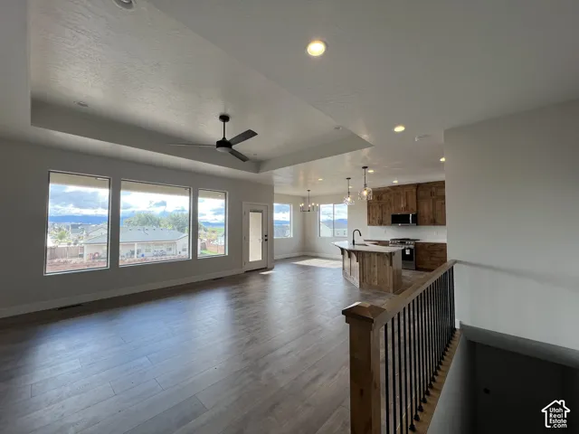 Open-concept interior featuring wood-finish flooring, a tray ceiling with a recessed ceiling fan, natural wood cabinetry, a center island with a built-in sink, and large windows providing mountain scenery