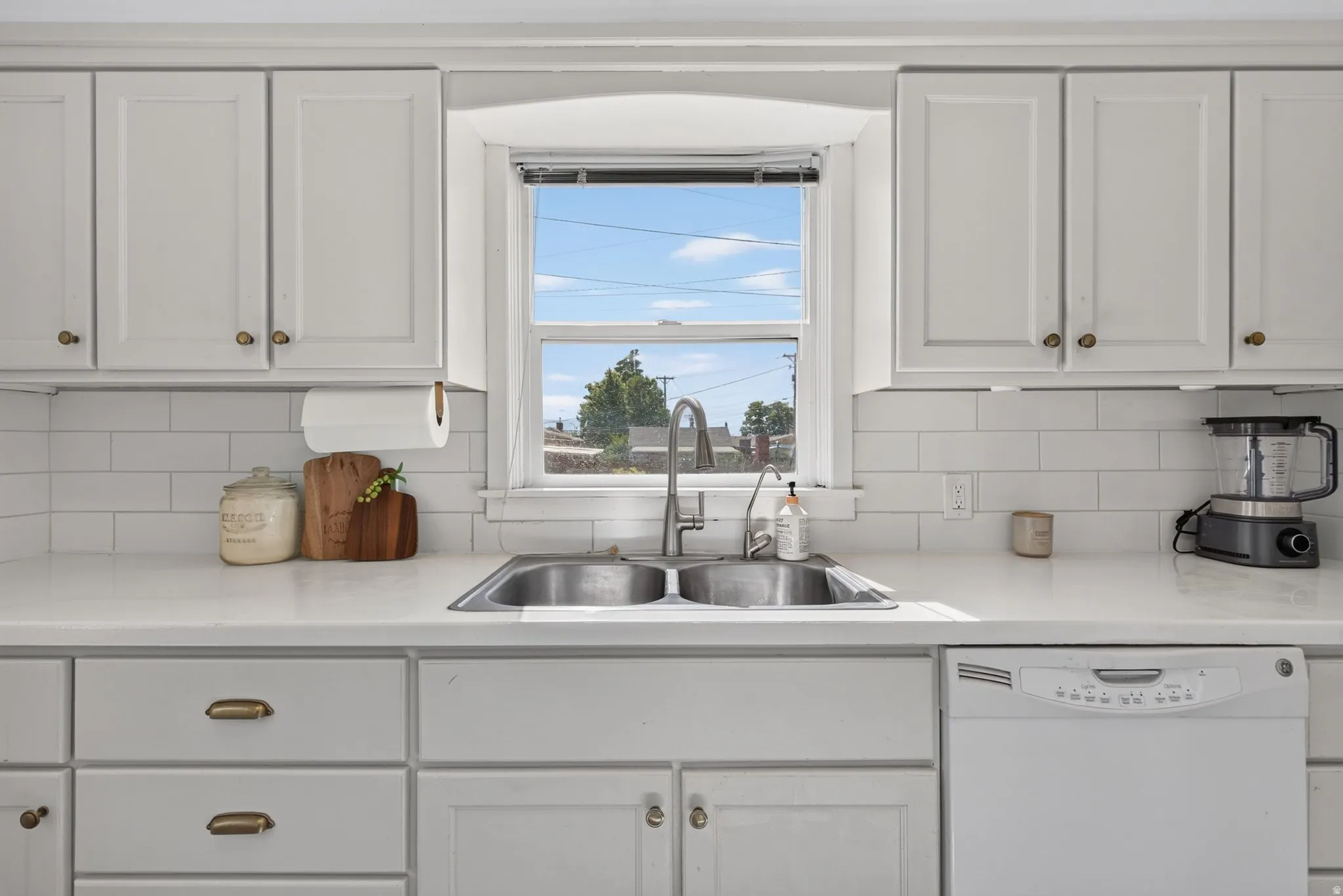 White cabinetry with brass hardware, a double basin stainless steel sink with gooseneck faucet, white subway tile backsplash, white countertops, and a built-in white dishwasher