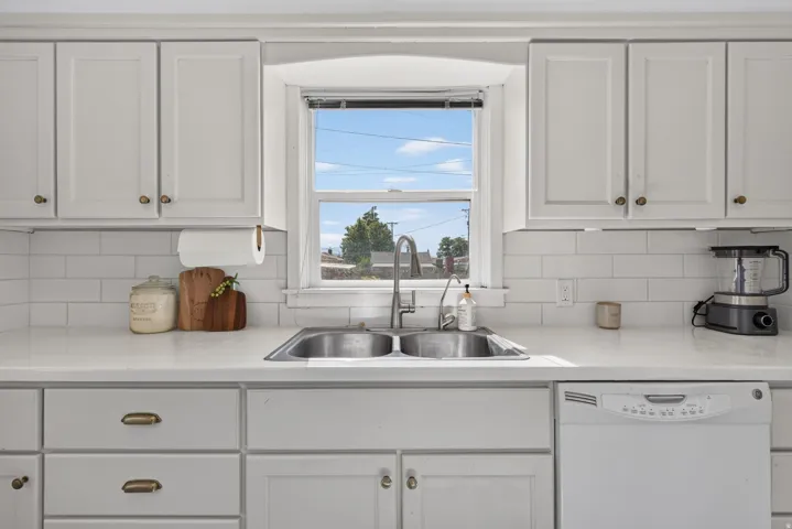 White cabinetry with brass hardware, a double basin stainless steel sink with gooseneck faucet, white subway tile backsplash, white countertops, and a built-in white dishwasher