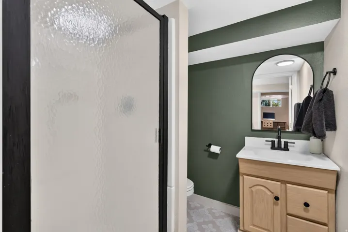Bathroom featuring a shower with a textured glass door and matte black frame, a wood-finish vanity with a white countertop, a black arched mirror, and a matte black faucet