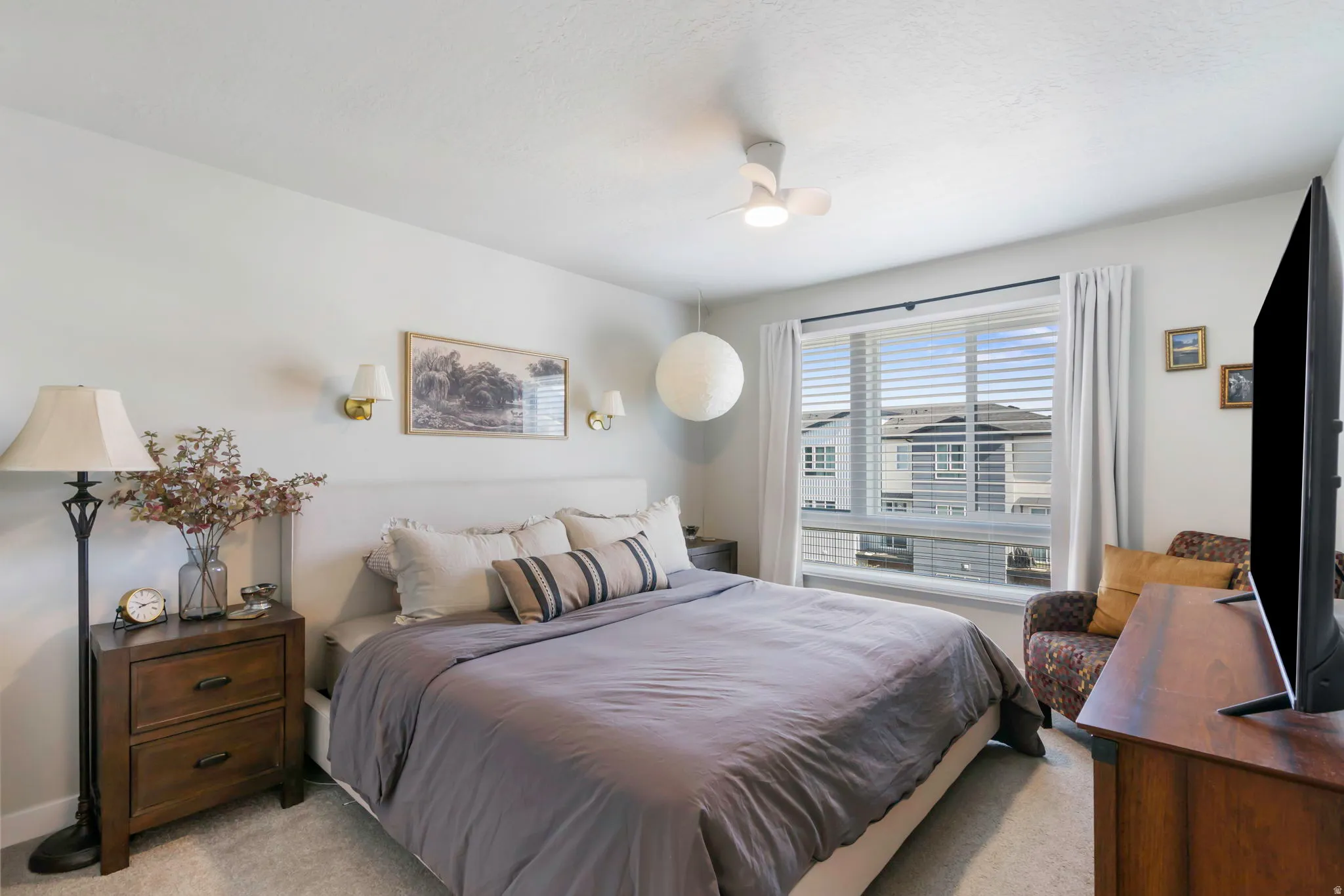 Spacious bedroom featuring light neutral wall paint, a large window with blinds and drapes, a ceiling fan, and wall-mounted sconces