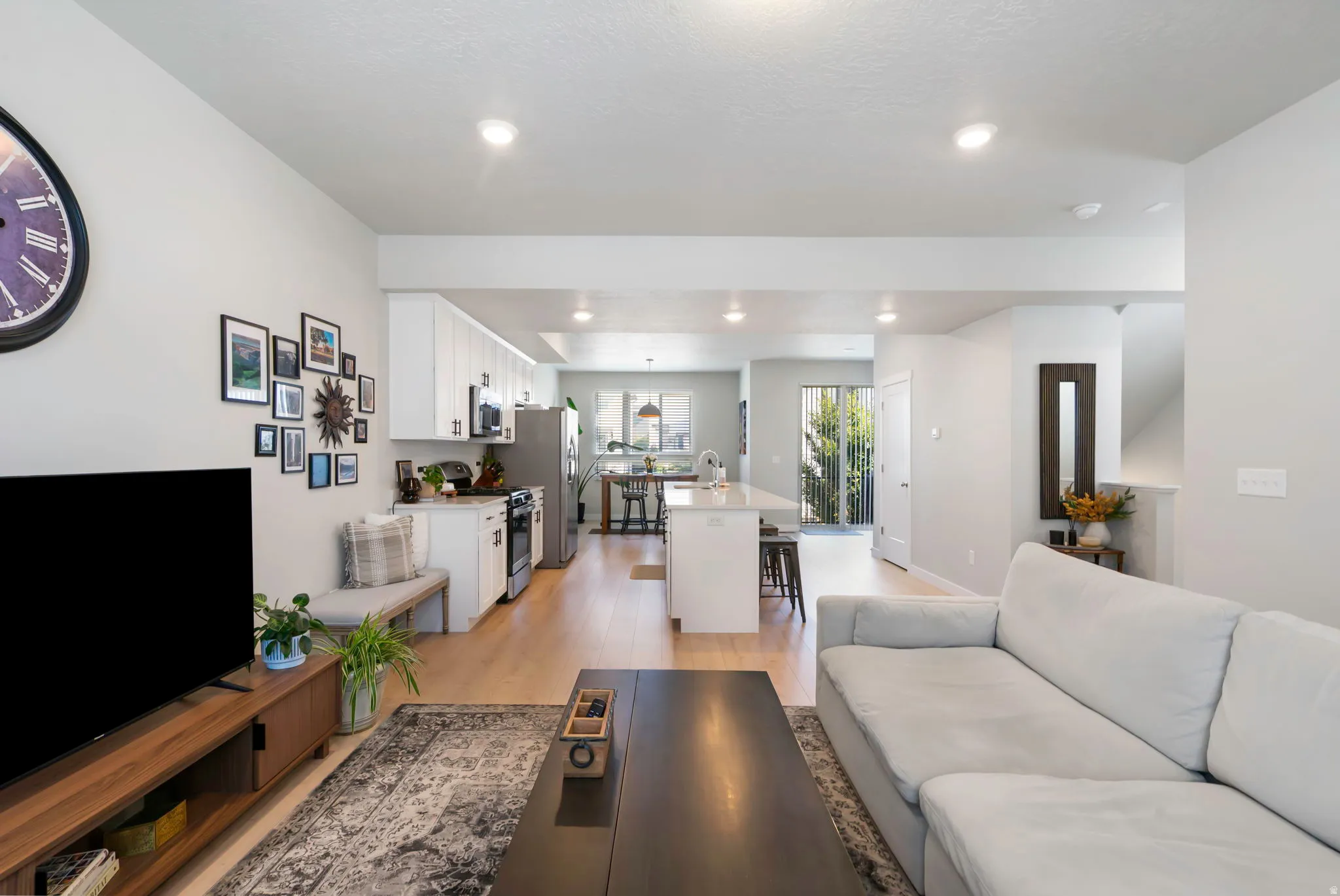 Open-concept living area featuring light wood-finish flooring, recessed lighting, a white kitchen with stainless steel appliances, and a kitchen island with seating