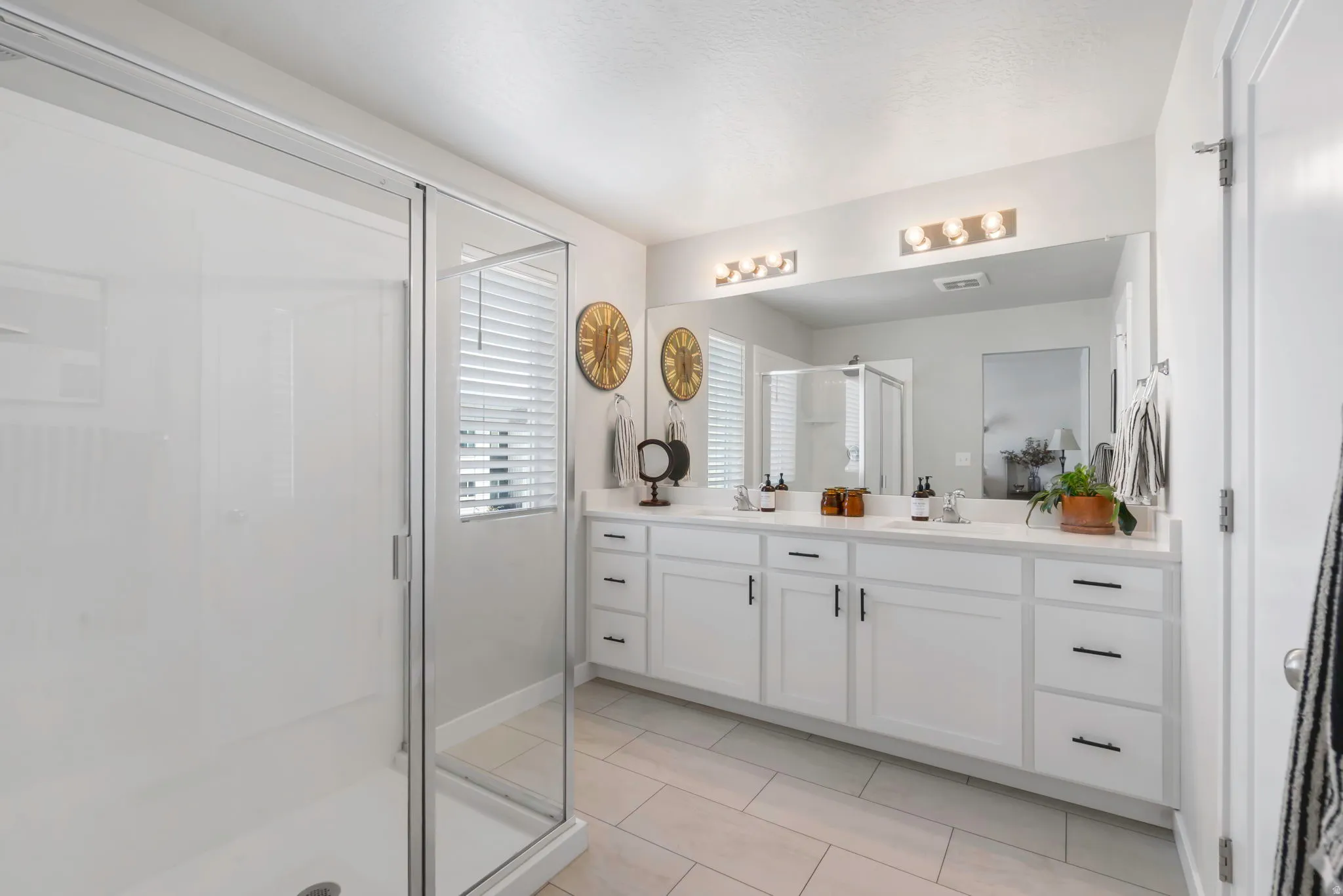 Spacious bathroom featuring a glass-enclosed shower, dual vanity with white cabinetry, matte black hardware, a large mirror, and tile flooring