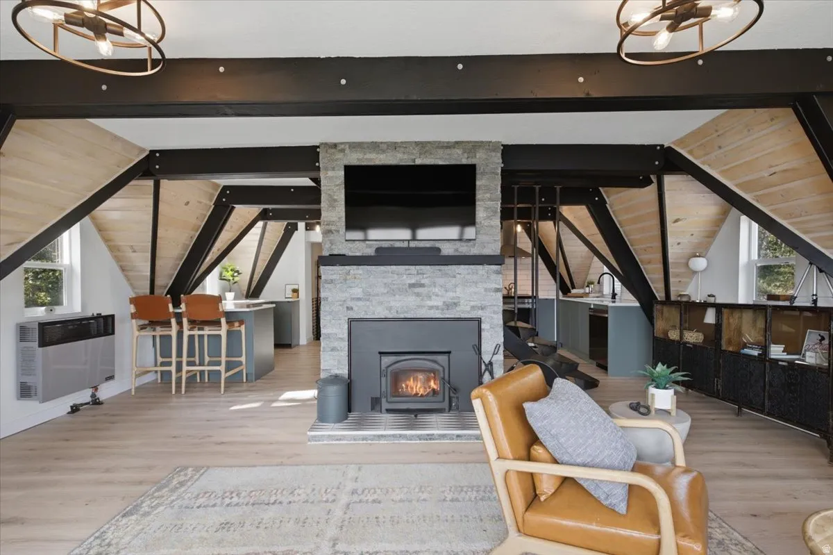 Living space featuring a stone fireplace with an integrated wood stove, exposed structural beams, wood-finish flooring, and two modern ceiling light fixtures