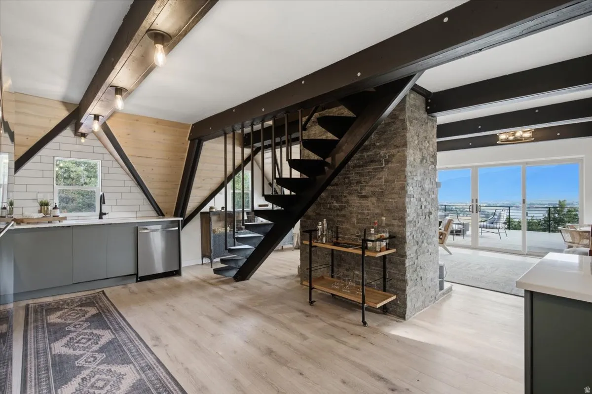 Open-concept interior featuring light wood-finish flooring, exposed ceiling beams, a modern spiral staircase, a kitchen with grey cabinetry and a stainless steel dishwasher, and sliding glass doors leading to an outdoor deck