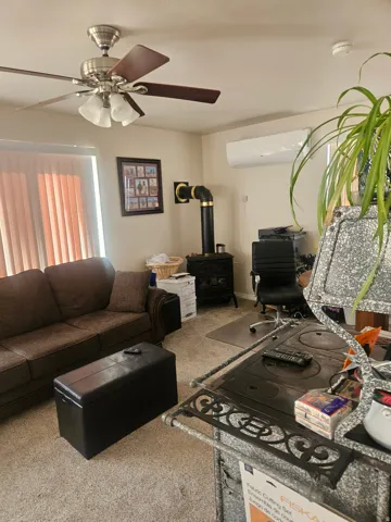 Carpeted living space featuring a ceiling fan with integrated lighting, wood-burning stove with a black pipe, and a wall-mounted ductless mini-split system