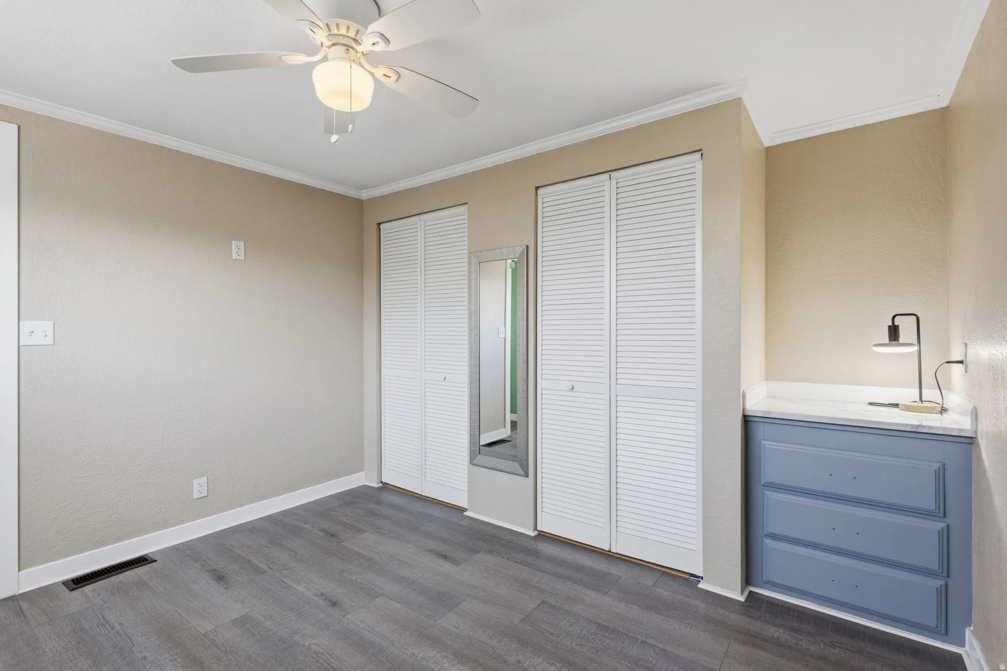 Bedroom featuring wood-finish flooring, two sets of louvered bi-fold closet doors, a ceiling fan with integrated lighting, and a built-in dresser with a white countertop