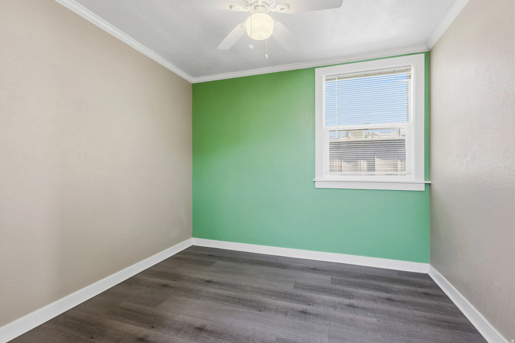 Room featuring wood-finish flooring, a vibrant accent wall, a window with blinds, a ceiling fan with integrated lighting, and white baseboards and crown molding