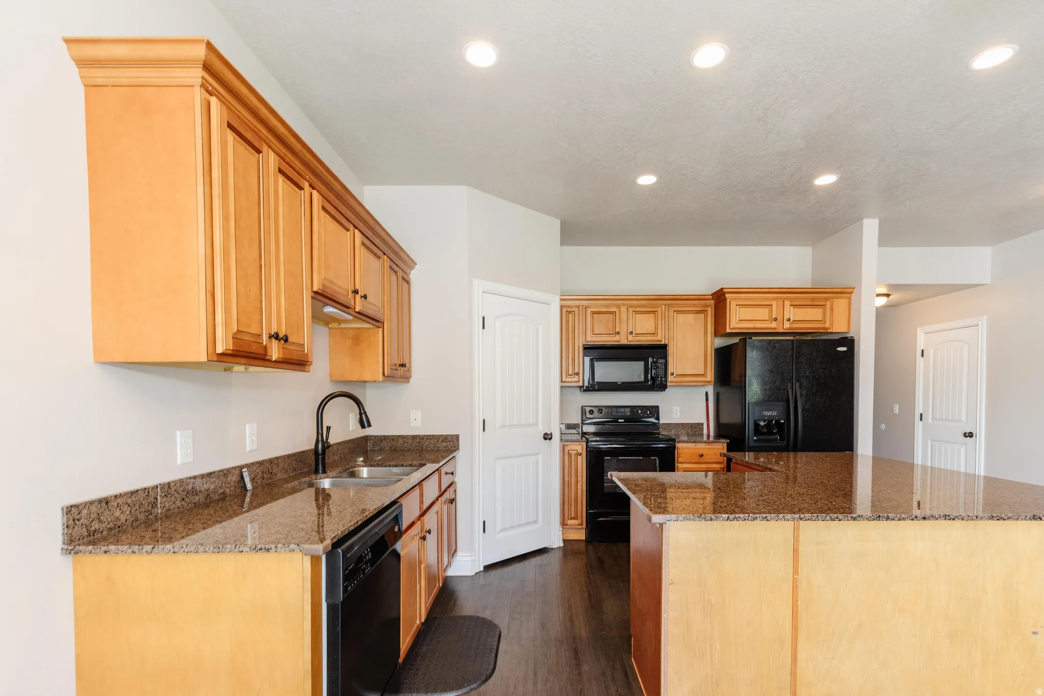 Kitchen featuring wood-finish cabinetry, granite-style countertops, a stainless steel double basin sink with a matte black faucet, recessed lighting, and black appliances