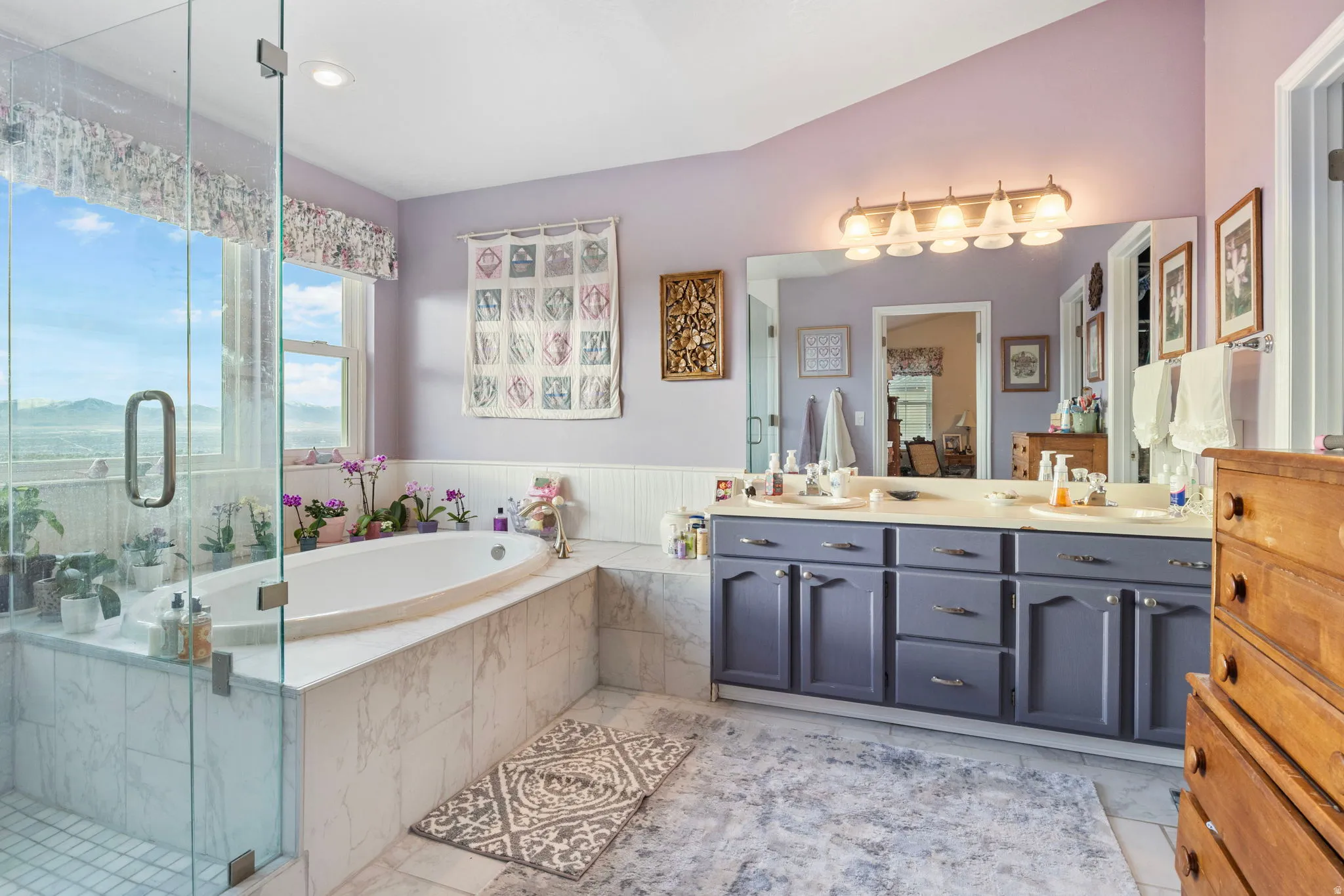 Spacious bathroom featuring a built-in soaking tub with marble-look surround, a separate glass-enclosed shower, a double vanity with white countertops, and expansive windows showcasing a mountain backdrop
