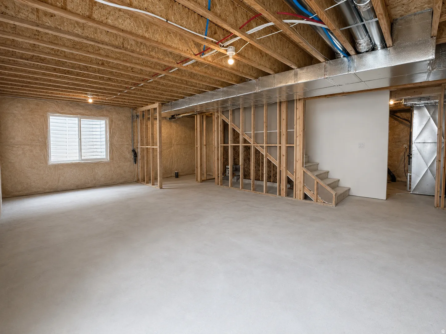 Unfinished basement featuring exposed wood joists, concrete flooring, a visible window, and framed walls with a partial staircase