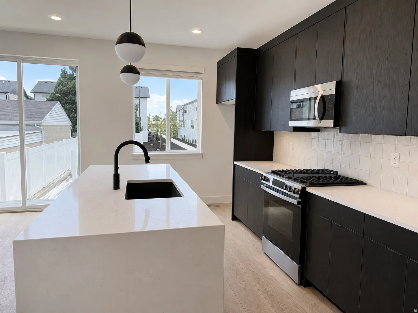 Kitchen featuring a waterfall-edge island with an undermount sink and matte black faucet, dark wood-finish cabinetry, stainless steel microwave, gas range, and light wood-finish flooring