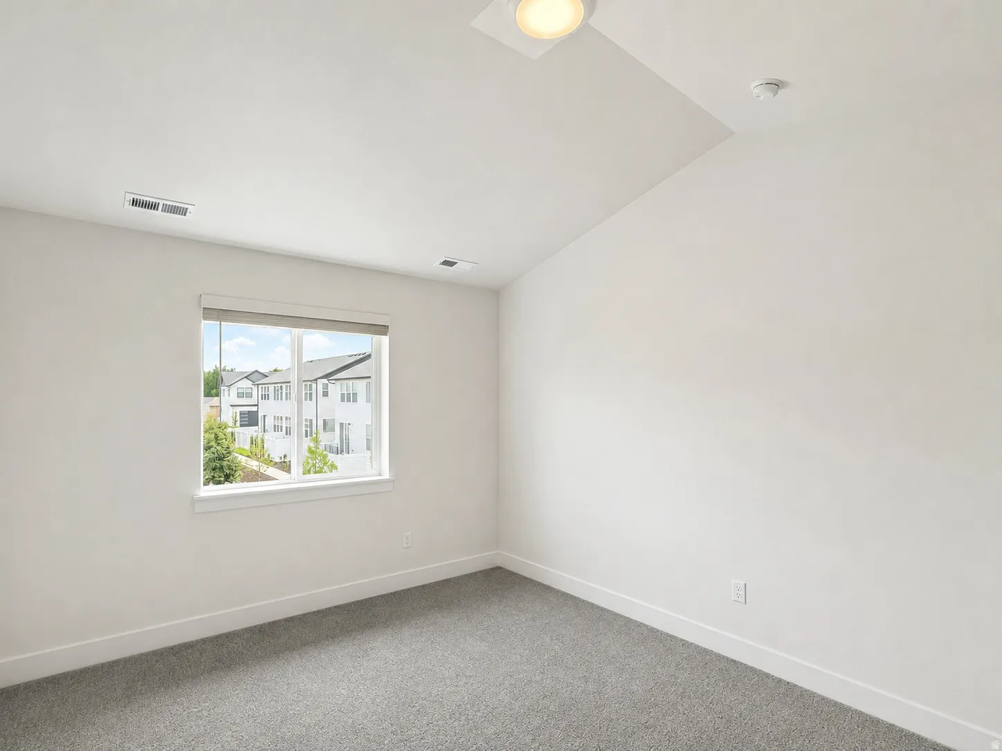 Bright room featuring neutral walls, gray carpet, a white framed window with roller shades, and a ceiling-mounted light fixture