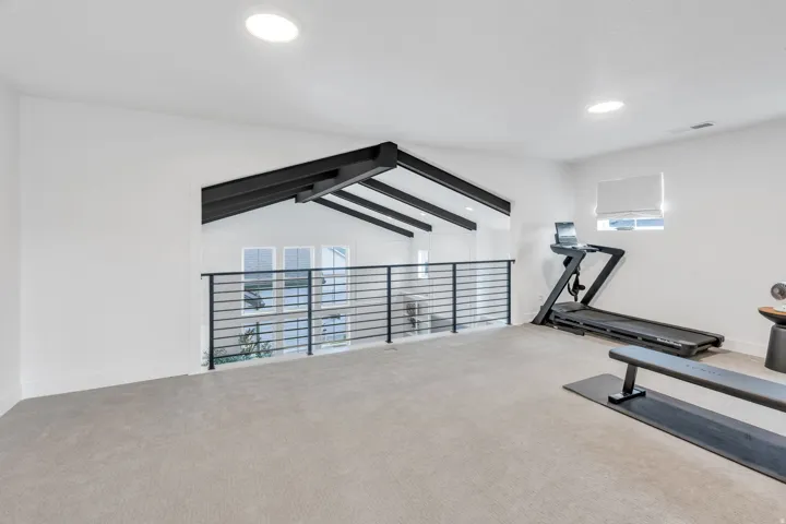 Open loft space featuring light-toned carpeting, recessed lighting, a modern horizontal bar railing, and exposed black ceiling beams