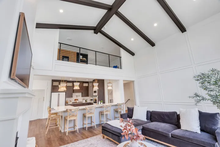 Vaulted ceiling with exposed wood beams, white paneled accent wall, open-concept kitchen with island seating, wood-finish flooring, and an upper-level loft with a black metal railing