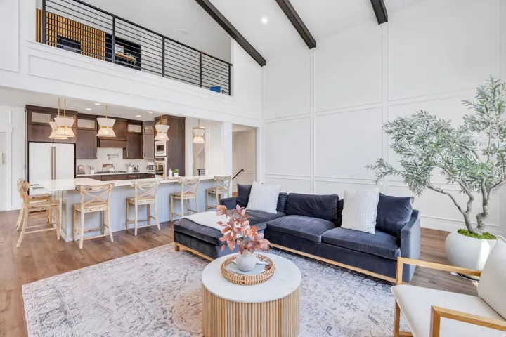 Open-concept living space featuring wood-finish flooring, a kitchen island with seating, pendant lighting, vaulted ceilings with exposed beams, and a second-story interior balcony with horizontal metal railings