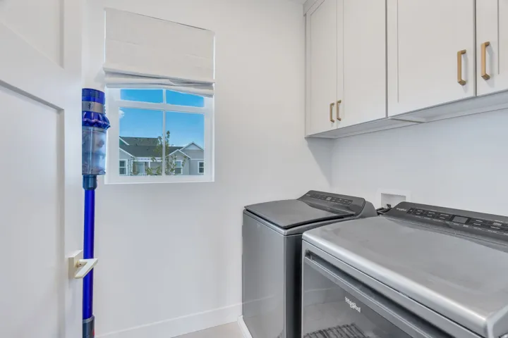 Laundry area featuring white cabinetry with gold-tone hardware, a window with a roman shade, and light-toned flooring