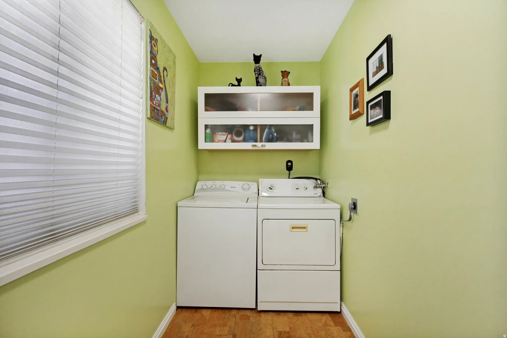 Dedicated laundry area featuring a window with blinds, upper white cabinetry with glass inserts, and wood-finish flooring