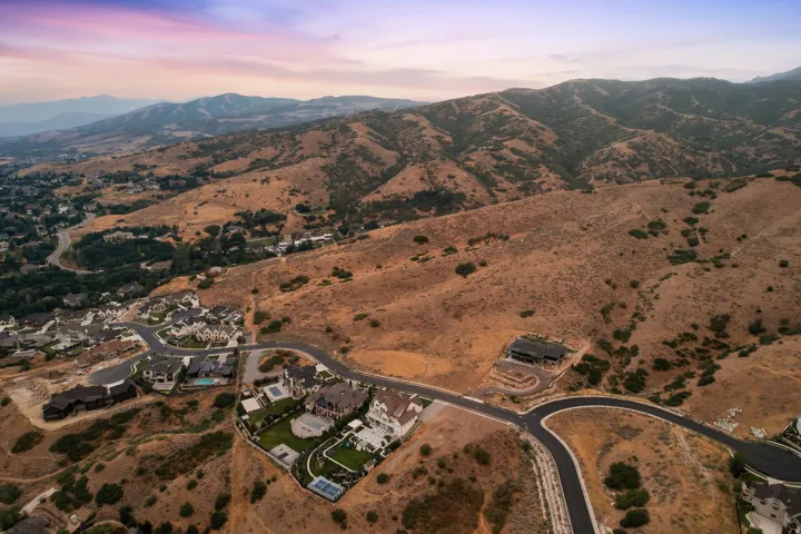 Expansive aerial view showcasing a developing residential area nestled against mountainous terrain, featuring multiple properties with diverse architectural styles, landscaped grounds, and paved roadways