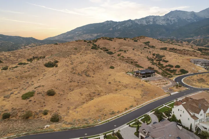 Expansive undeveloped land parcel with mountainous backdrop, featuring paved roadways and existing residential structures nearby