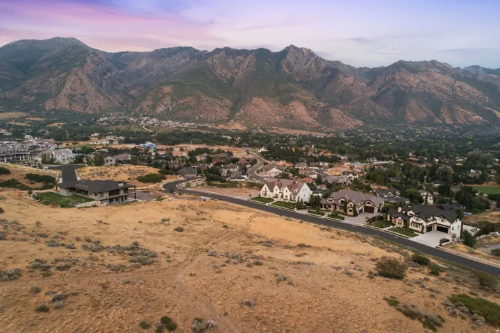 Elevated residential area with mountain backdrop, featuring a modern, multi-level home with a dark roof and a series of newly constructed, diverse architectural style residences along a paved road