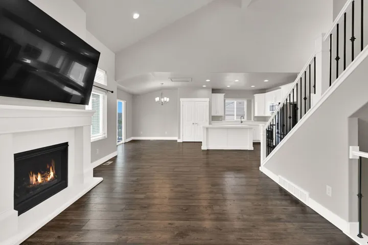 Spacious open-concept interior featuring wood-finish flooring, a contemporary fireplace with white mantel, vaulted ceilings, and a kitchen with white cabinetry and an island