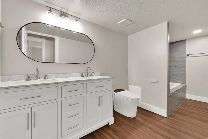 Spacious bathroom featuring a double vanity with white cabinetry and a light-toned countertop, wood-finish flooring, a wall-mounted oval mirror, modern vanity lighting, and a built-in tub with a tiled surround