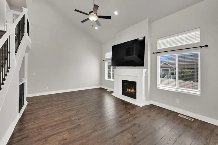 Living room featuring vaulted ceilings, wood-finish flooring, a contemporary fireplace with white mantel, a ceiling fan with integrated lighting, and a staircase with wrought iron balusters