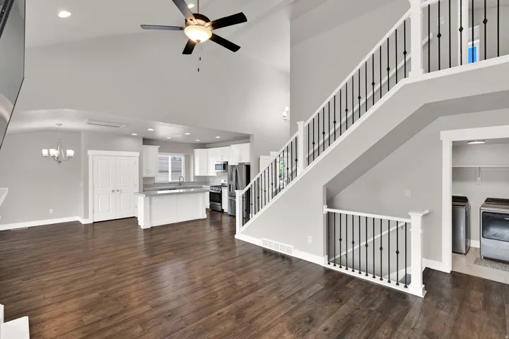 Vaulted ceiling with a ceiling fan, hardwood-finish flooring, white kitchen cabinetry, stainless steel appliances, and an open staircase with black metal balusters