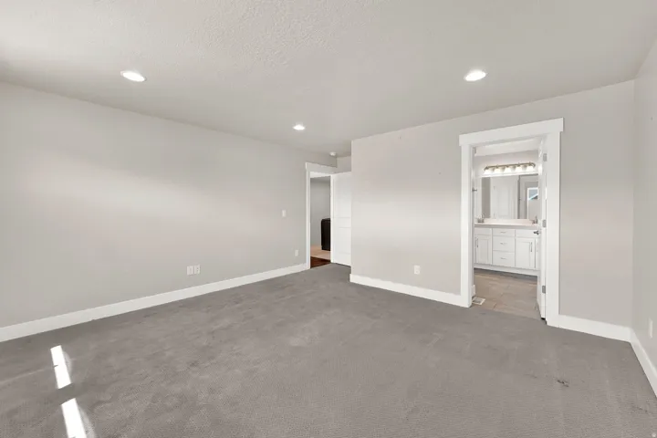 Spacious carpeted room featuring recessed lighting and a doorway revealing a bathroom with a white vanity, dual mirrors, and contemporary lighting fixtures