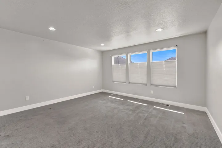 Bright primary bedroom featuring three large windows with cellular shades, recessed lighting, neutral wall paint, white baseboards, and gray carpeting